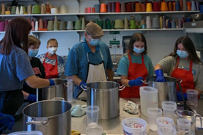 Ceramics art students wearing gloves and goggles gather around a table of pots and containers. They are learning the process of bath agitation and the whole dye process during the second installment of a three-part workshop on Furoshiki and Shibori. 