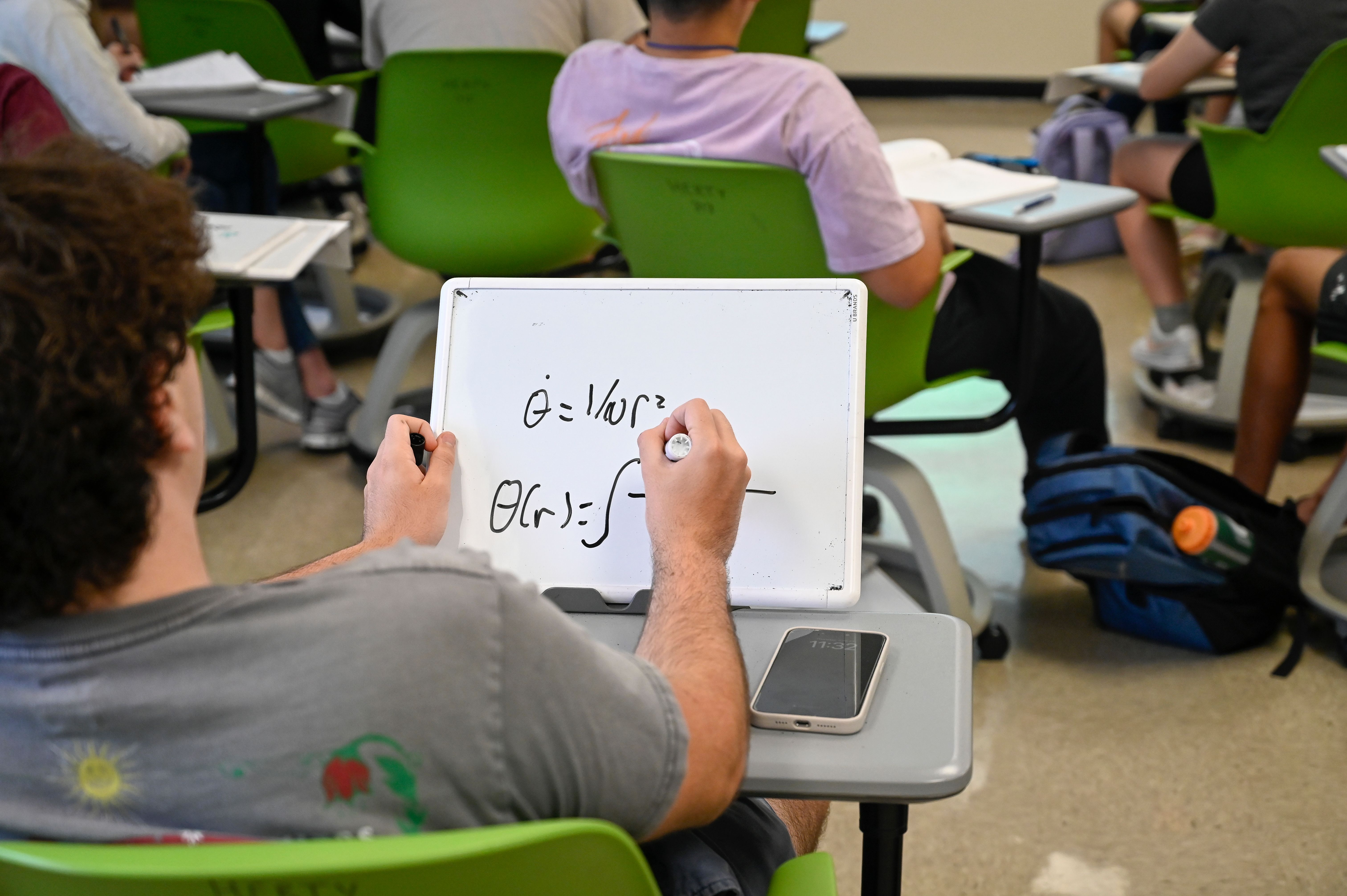 Over shoulder view of a SSP student drawing an equation on a wipey board.