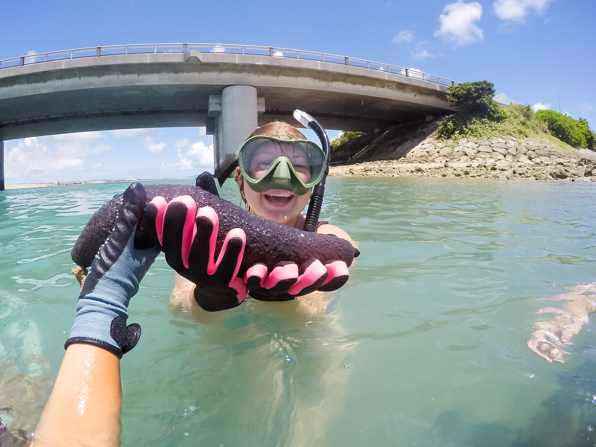 A student in goggles lifts a sea cucumber out of the sea, smiling.